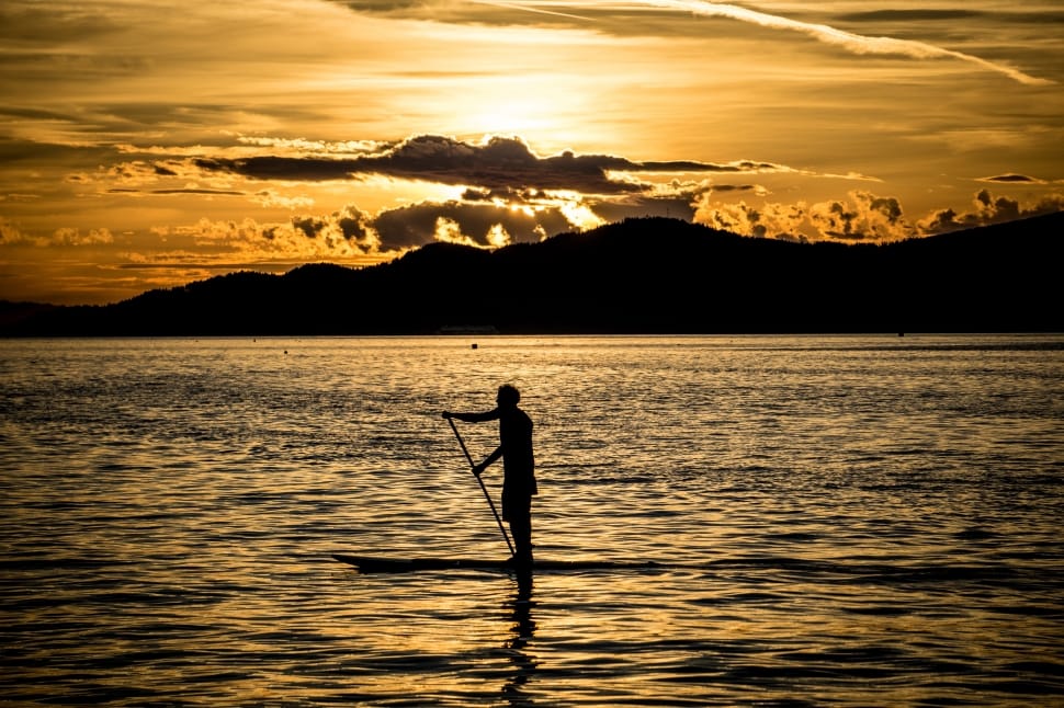 silhouette of a person standing on top of boat holding stick with mountains preview