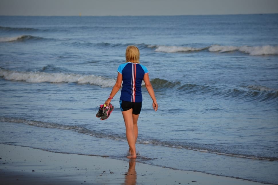 woman in blue and black shirt holding sneaker walking near seashore during daytime preview