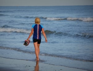 woman in blue and black shirt holding sneaker walking near seashore during daytime thumbnail