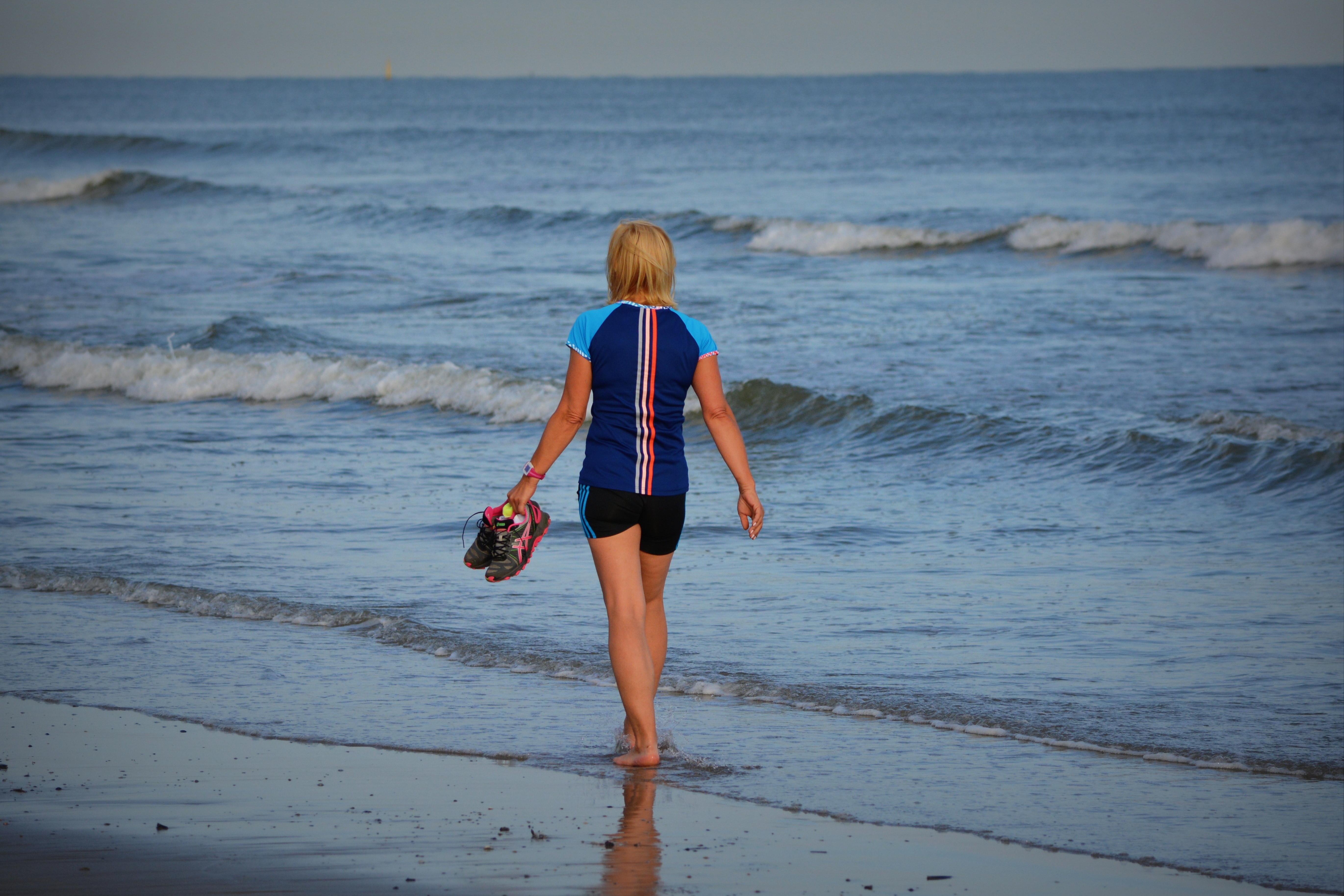 woman in blue and black shirt holding sneaker walking near seashore during daytime