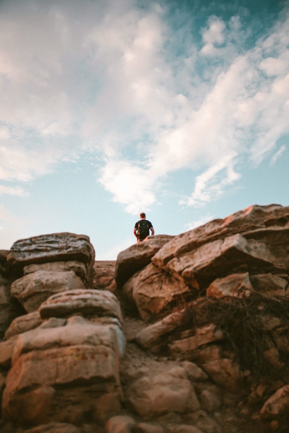 man in black t shirt and backpack standing on brown rock formation under white clouds and blue sky preview