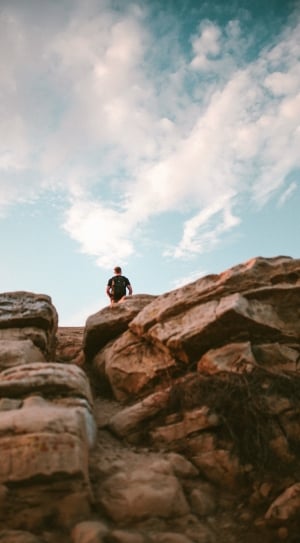 man in black t shirt and backpack standing on brown rock formation under white clouds and blue sky thumbnail