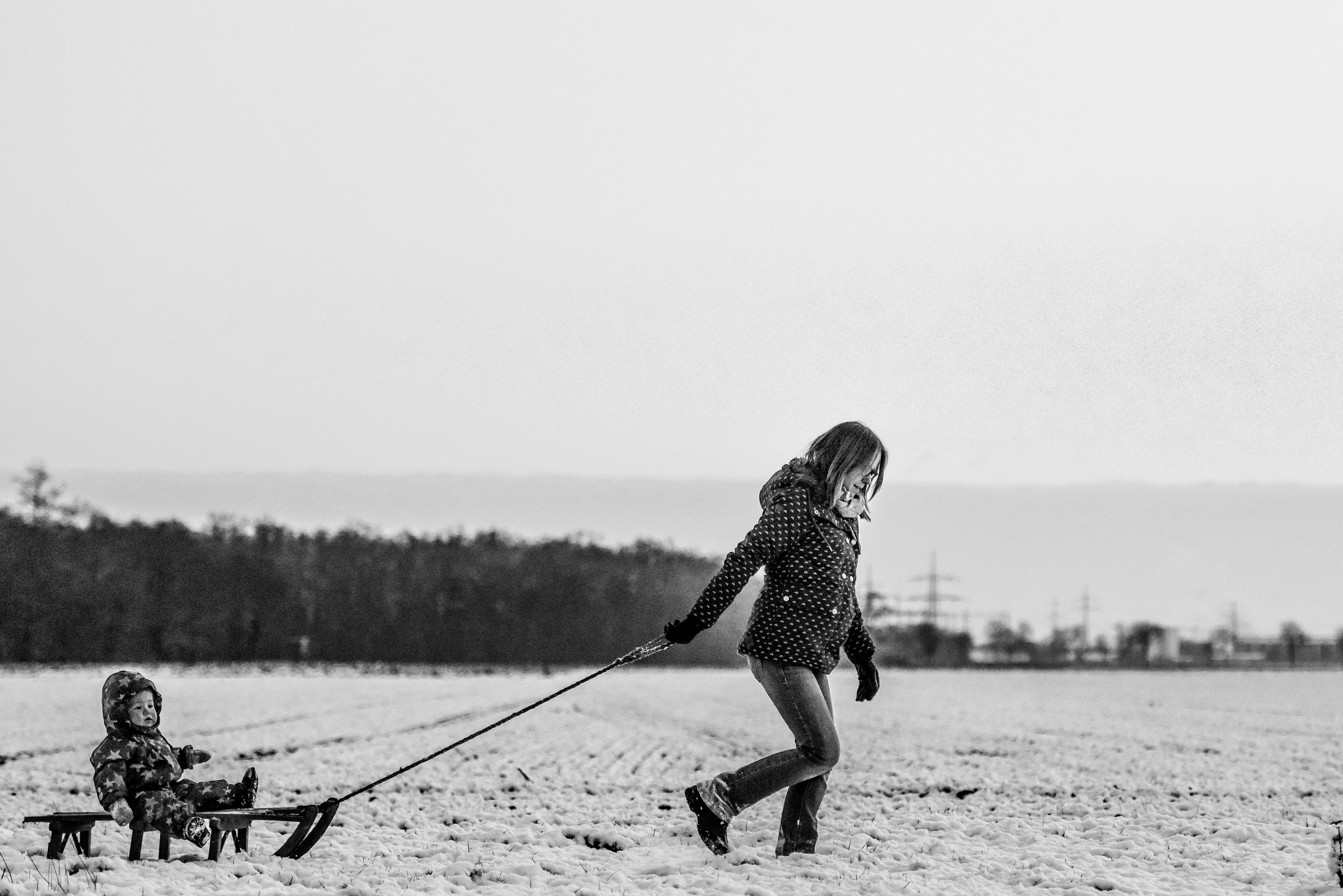 graycale photography of woman pulling child in cart on snow covered ground beside forest