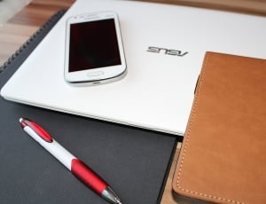 white and red click pen beside brown leather case and white android smartphone on white Asus laptop thumbnail
