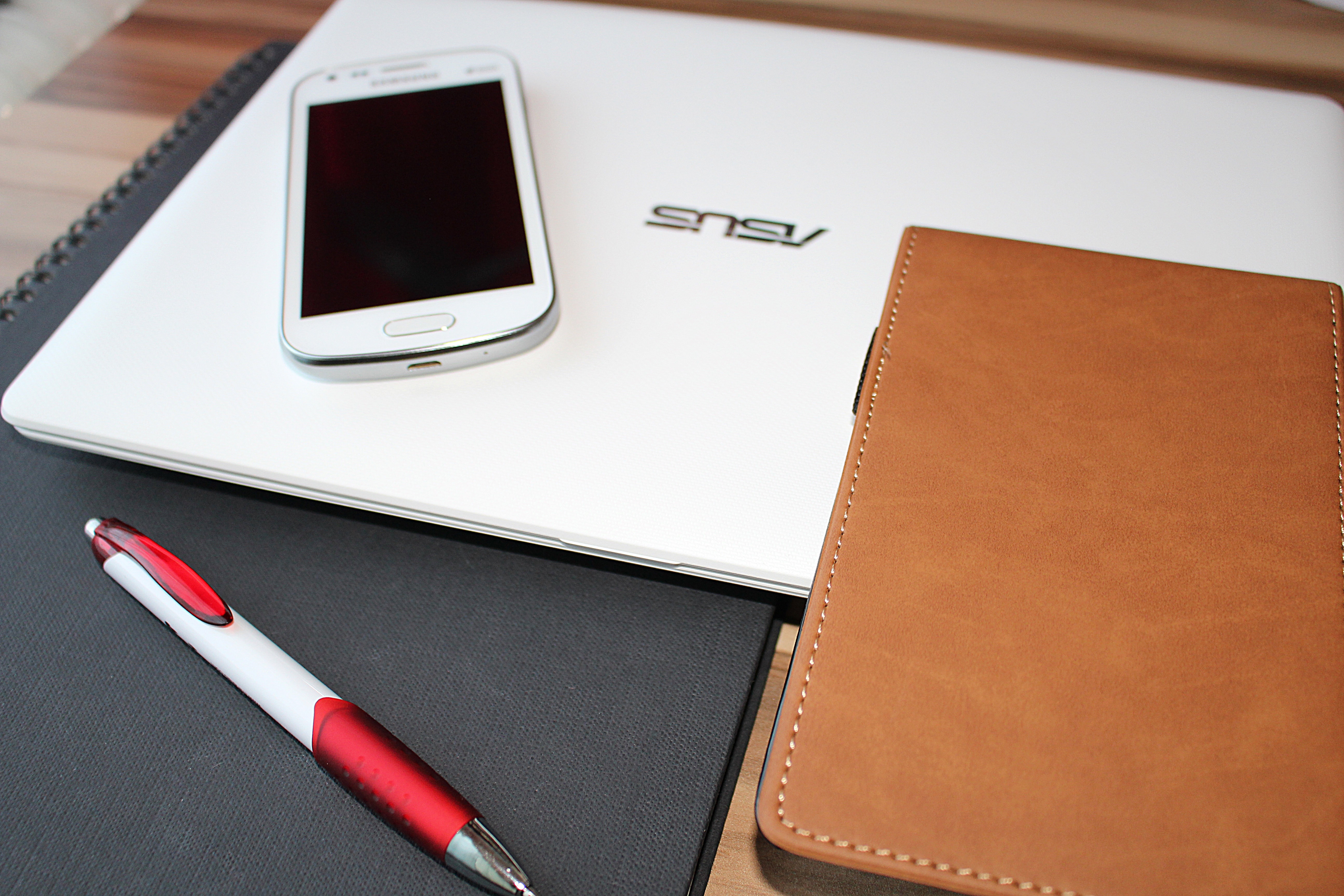 white and red click pen beside brown leather case and white android smartphone on white Asus laptop