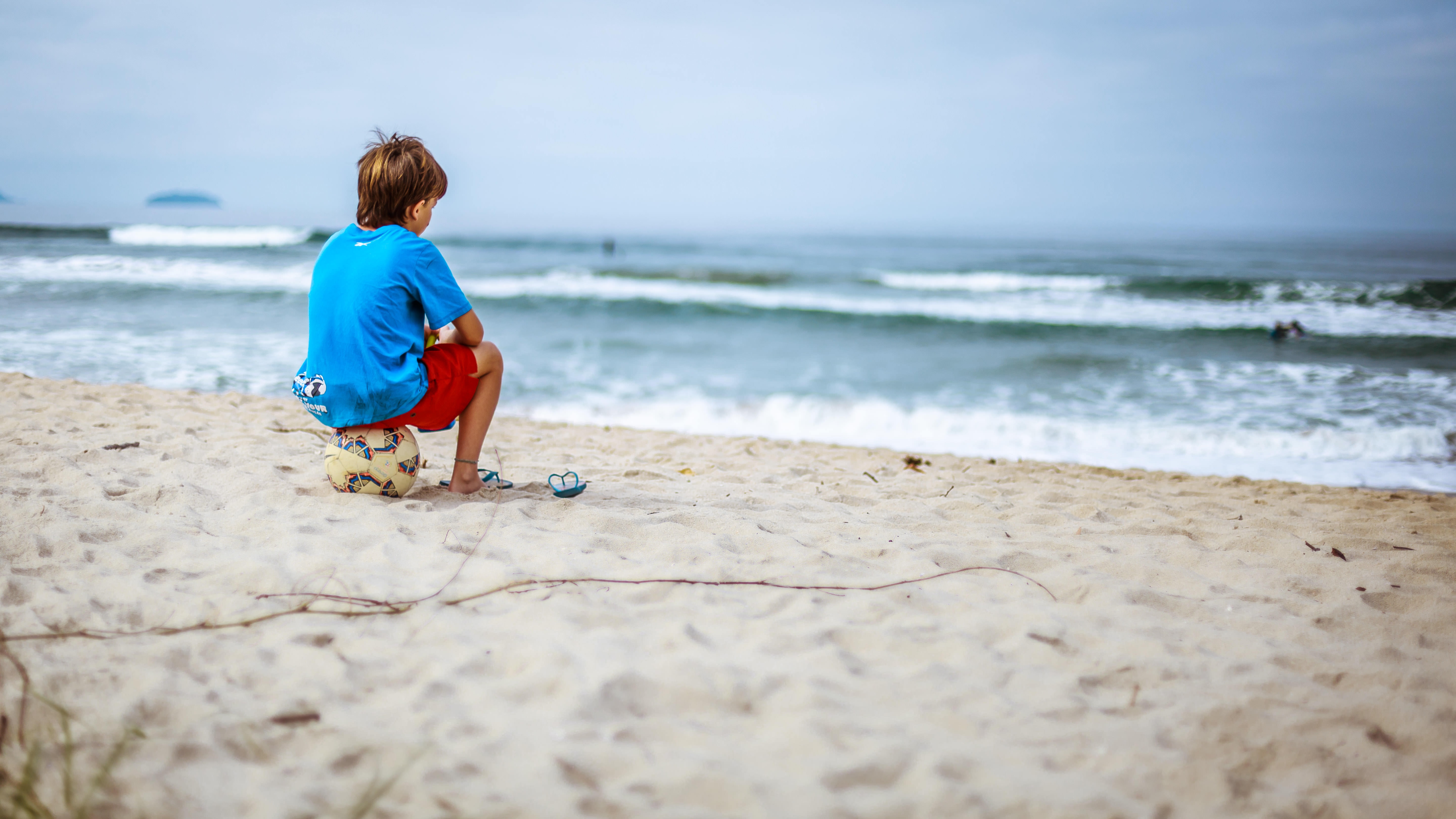 boy sitting on soccer ball by the beach while watching the sea waves during cloudy skies