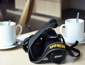 black Nikon DSLR camera with two white ceramic mug in saucers thumbnail