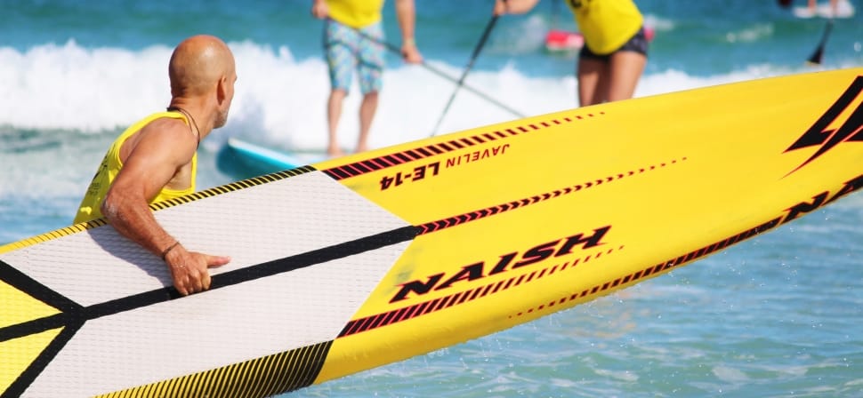 man wearing yellow tank top holding white and yellow naish surfboard facing body of water during daytime preview