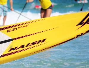 man wearing yellow tank top holding white and yellow naish surfboard facing body of water during daytime thumbnail