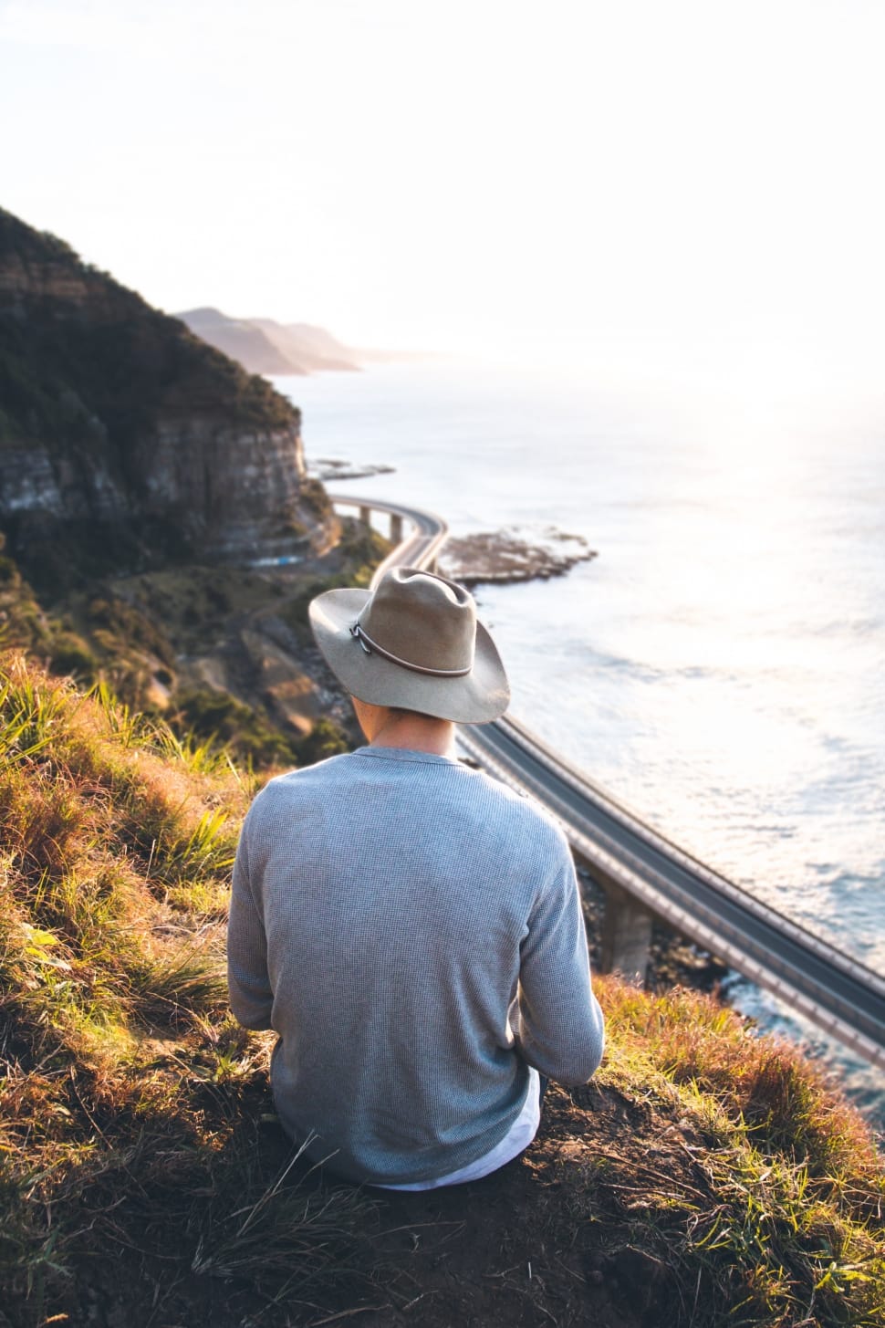 man sitting wearing gray sweater and gray cowboy hat on top of hill over viewing body of water preview