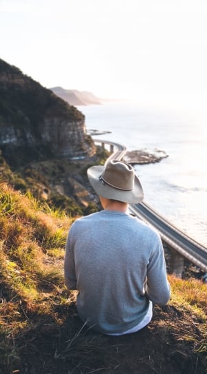 man sitting wearing gray sweater and gray cowboy hat on top of hill over viewing body of water thumbnail