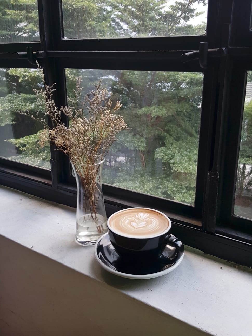 black ceramic mug filled with cappuccino coffee with saucer beside a clear glass flower vase and black wooden framed window preview