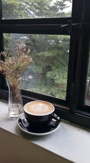 black ceramic mug filled with cappuccino coffee with saucer beside a clear glass flower vase and black wooden framed window thumbnail