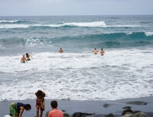 5 persons swimming at beach near 3 persons at gray seashore during daytime thumbnail