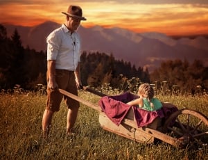 girl riding on brown wooden wheelbarrow carrying by man during sunset thumbnail