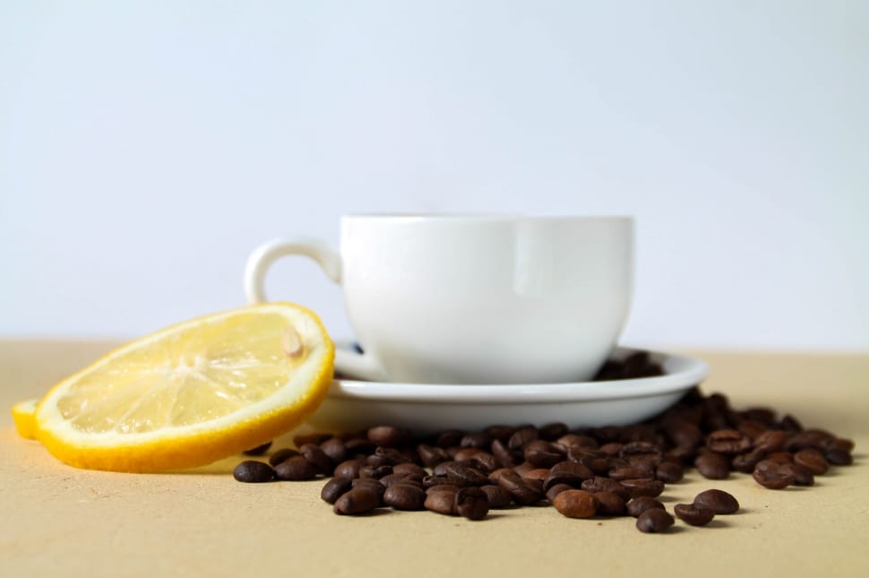 white ceramic cup on saucer surrounded by coffee beans beside slice of lemon preview