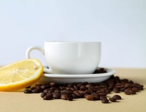 white ceramic cup on saucer surrounded by coffee beans beside slice of lemon thumbnail