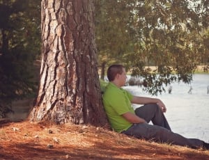man in green collar shirt sitting on lining on tree watching body of water during daytime thumbnail