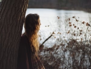 woman wearing brown long sleeve dress leaning on a brown tree trunk during daytime thumbnail