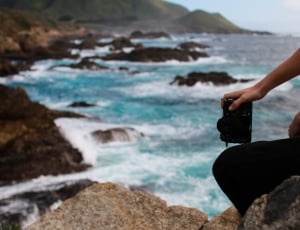 person holding camera sitting in rock near body of water thumbnail
