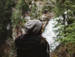 woman in black jacket sitting near waterfall surrounding by tress thumbnail