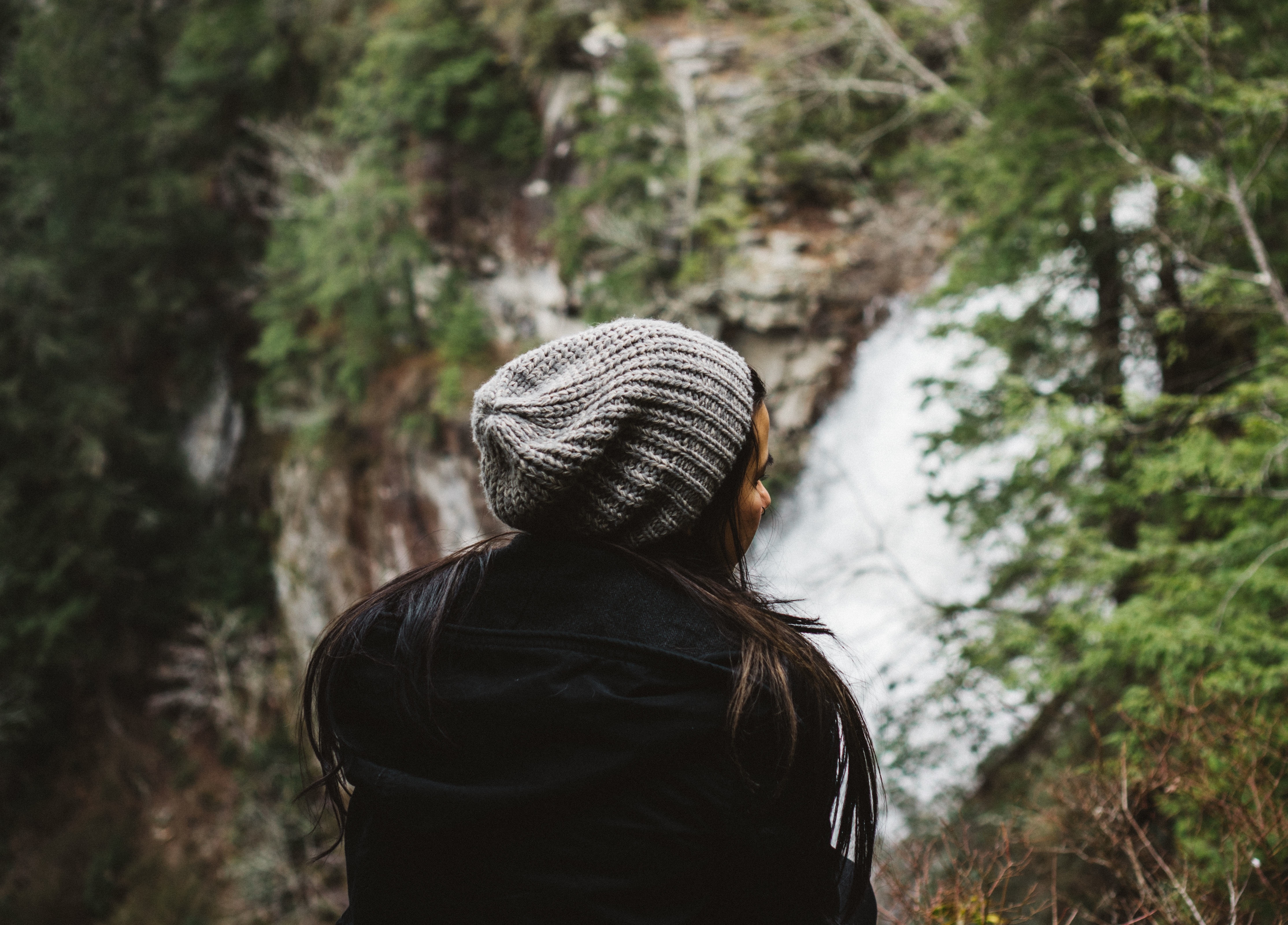 woman in black jacket sitting near waterfall surrounding by tress
