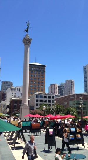 white column statue surrounded by patio umbrellas and buildings under blue sky during daytime thumbnail