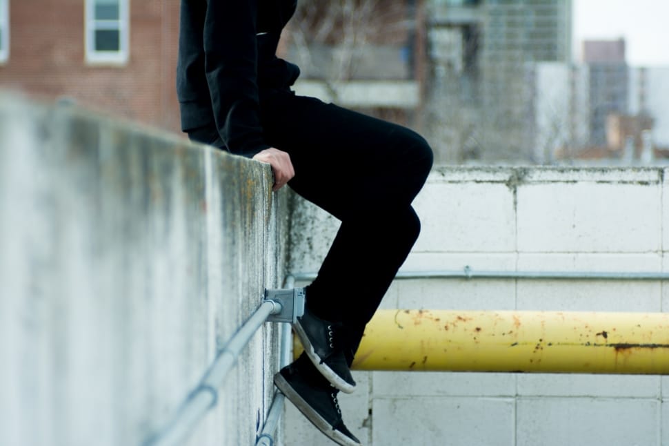 man wearing black pants and black shoes sitting on grey concrete wall with grey pipes preview