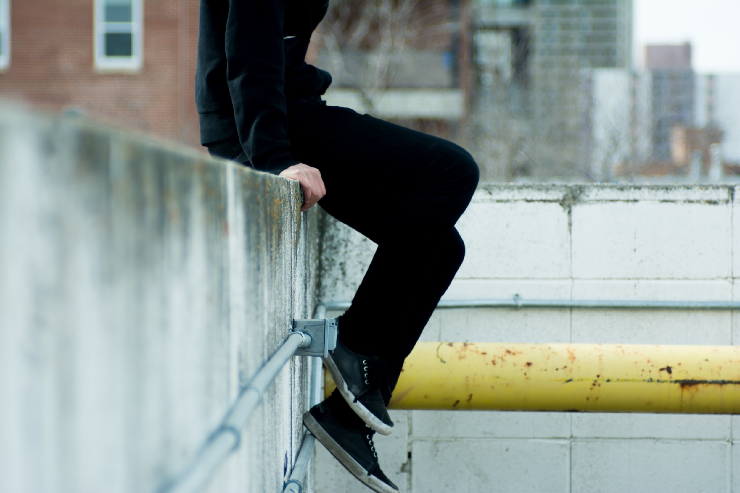 man wearing black pants and black shoes sitting on grey concrete wall with grey pipes