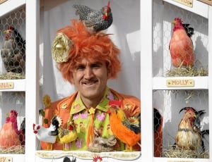 man in orange formal suit jacket surrounded of roosters and chicks toys thumbnail