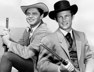 grayscale photo of two men with hats holding guns and smiling thumbnail