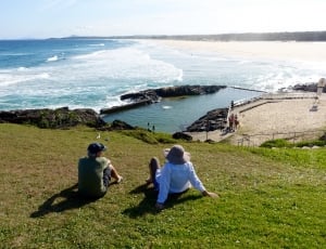 2 person sitting on green grass field and seashore during daytime thumbnail