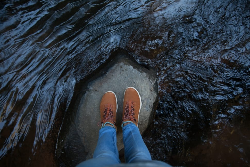person in brown shoes and blue jeans standing on rock in the middle of body of water preview