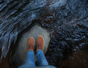 person in brown shoes and blue jeans standing on rock in the middle of body of water thumbnail