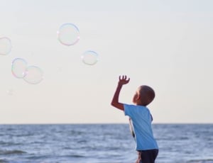 boy playing bubbles near body of water during daytime thumbnail