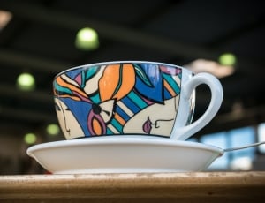 white, blue, pink, and brown ceramic coffee cup and saucer set on brown wooden table thumbnail