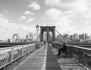 grayscale photo of man sitting on bench on bridge thumbnail