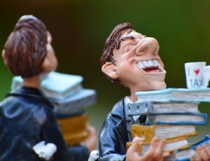 man in black coat holding pile of books figurines thumbnail