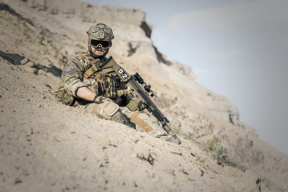 man in green and brown army suit holding rifle sitting on grey sand during day time preview