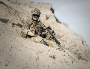 man in green and brown army suit holding rifle sitting on grey sand during day time thumbnail