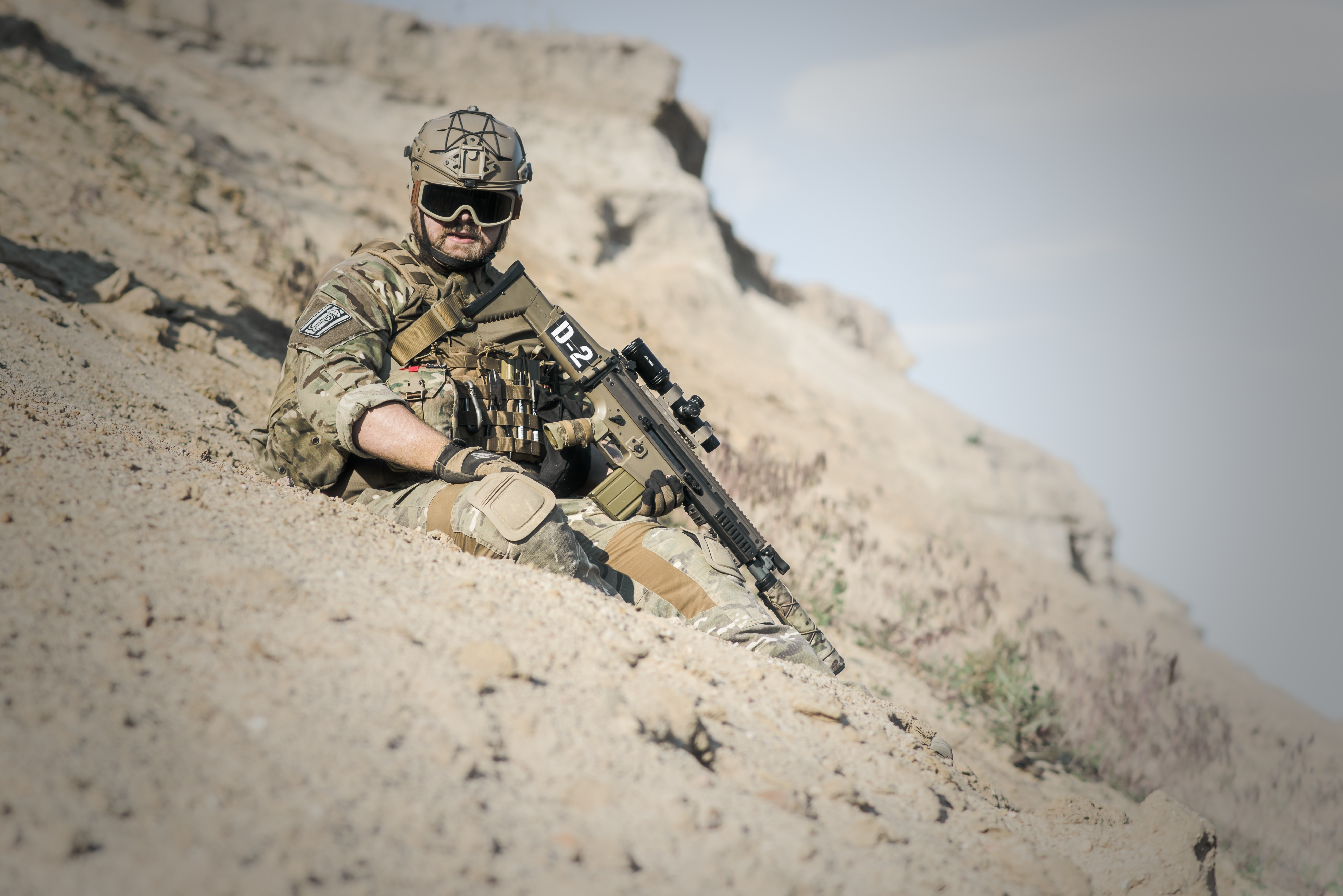 man in green and brown army suit holding rifle sitting on grey sand during day time
