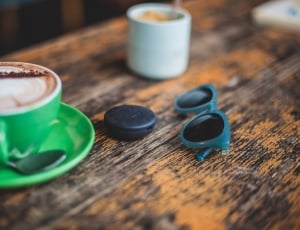 blue frame black lens sunglasses beside black round plastic container and green ceramic teacup filled with cappuccino thumbnail