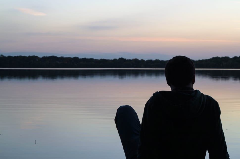 silhouette of man sitting beside large body of water during sunset preview
