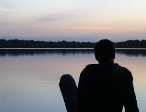 silhouette of man sitting beside large body of water during sunset thumbnail