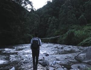 person with backpack standing on river in middle of forest during daytime thumbnail