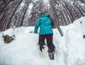 person in teal winter jacket and black pants carrying black and green snowboard walking on snow covered field in forest during daytime thumbnail