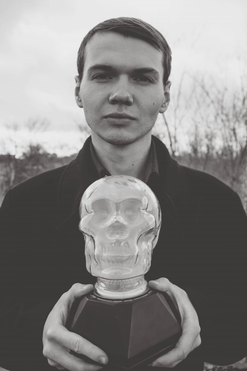 grayscale photo of a man in jacket holding a glass skull figurine during daytime preview