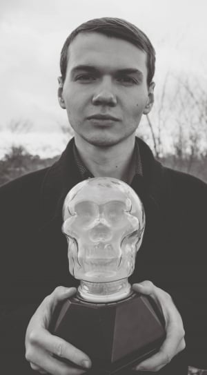 grayscale photo of a man in jacket holding a glass skull figurine during daytime thumbnail