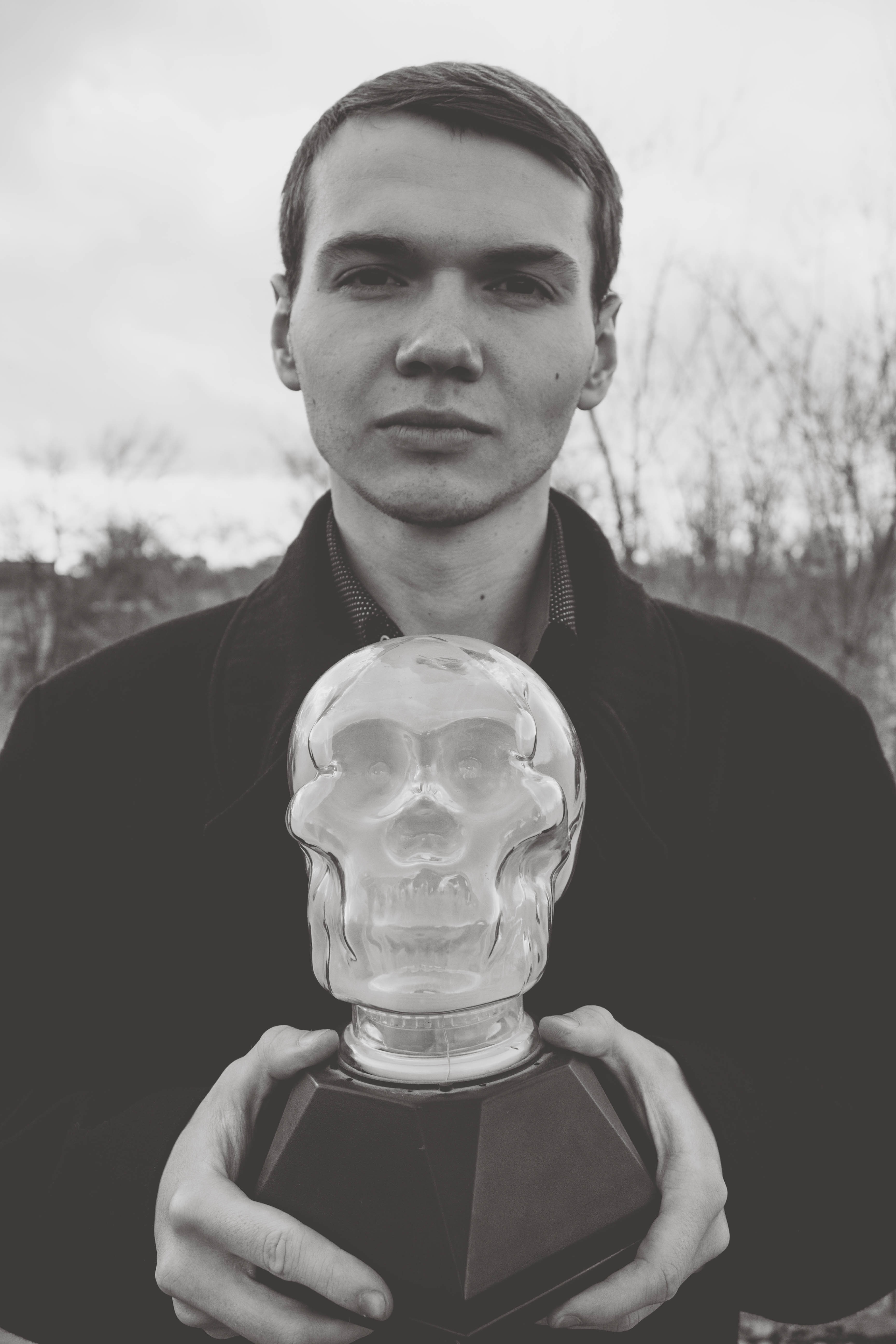grayscale photo of a man in jacket holding a glass skull figurine during daytime