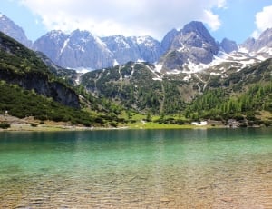 body of water near mountain peak and trees under white clouds thumbnail